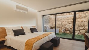 A modern bedroom with a large bed, a stone wall visible through the window, and a wooden floor. - Empreendimento Serra (Covilhã)
