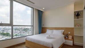 A hotel room with a large window, a wooden bed, a bedside table with a vase, and a shelf with decorative items.