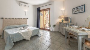 A bedroom with a bed, a desk, and a chair. There is a window with curtains, a picture on the wall, and a stone wall outside.