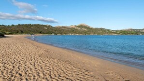 A sandy beach with clear blue water and a hill in the background.