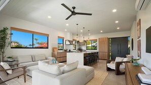 A modern living area with a sofa, coffee table, and dining table. There is a kitchen with a central island and bar stools. The room has large windows and a ceiling fan. - Modern Kauaʻi Luxury Home With Ohana, Pool, and Spa in Kukuiʻula (Koloa)
