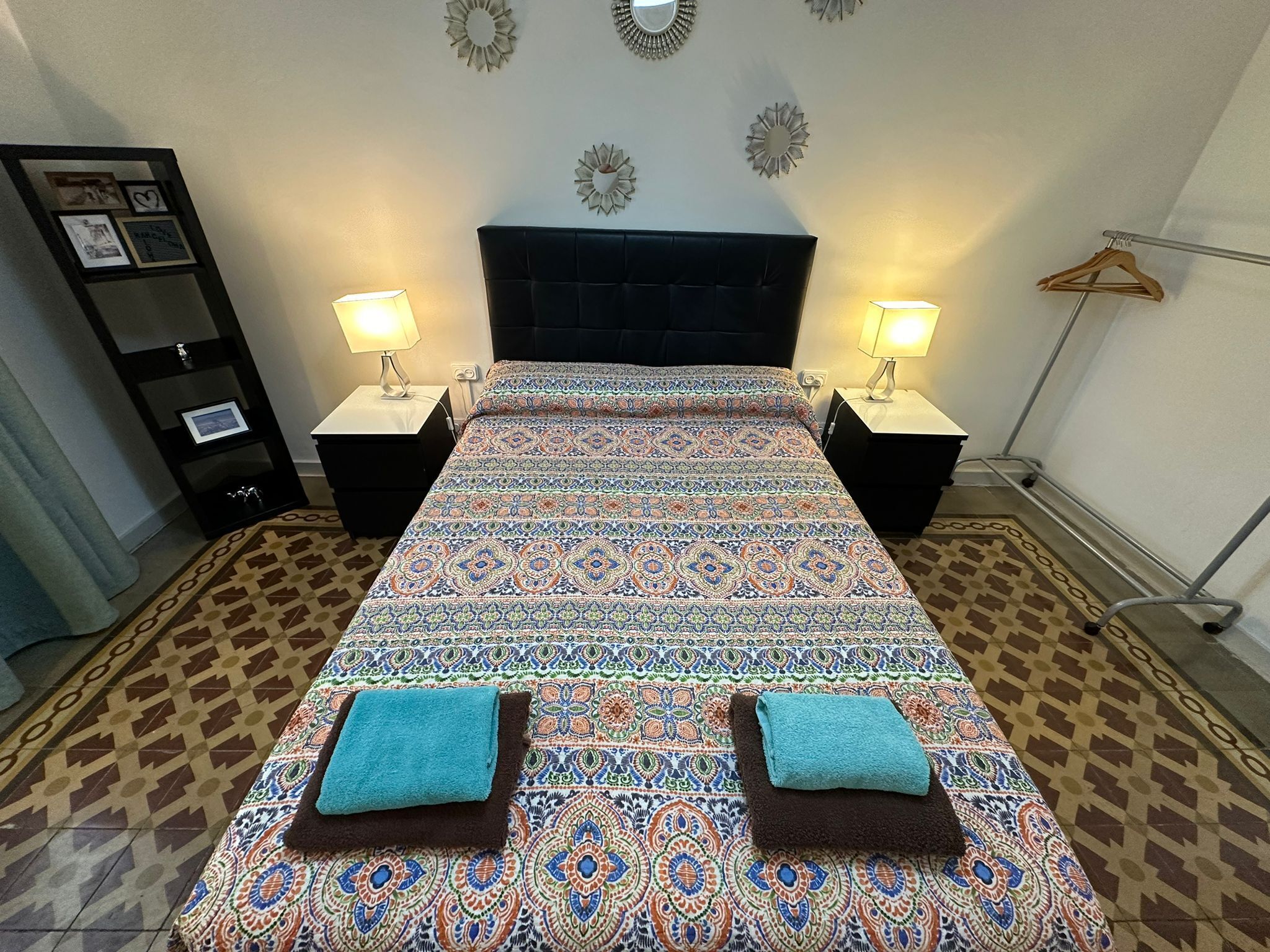 Una cama bien hecha con ropa de cama estampada, dos mesitas de noche con lámparas, un estante en la pared con fotografías y un suelo estampado.