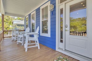 A covered porch with white chairs and a table, a blue exterior wall, and a white door with a window.