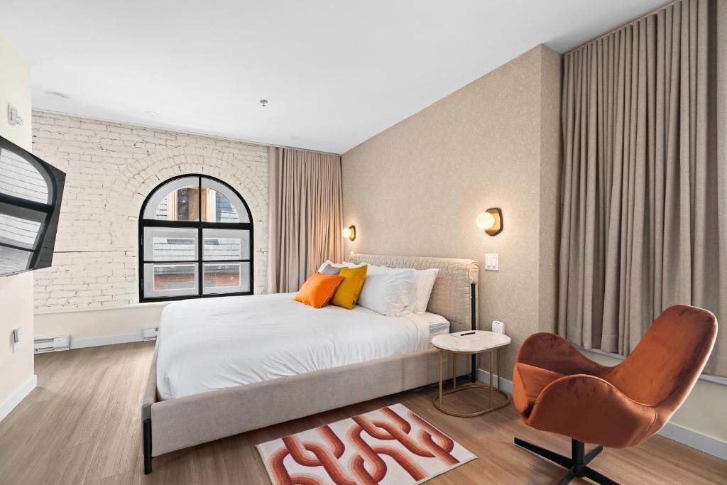 A modern bedroom with a large bed, a small side table, a chair, and a brick wall.