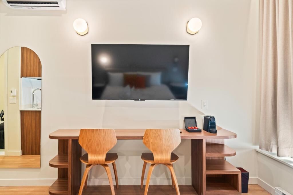 Ein modernes Hotelzimmer mit Flachbildfernseher an der Wand, einem Holztisch mit zwei Stühlen und einem kleinen Tisch mit Telefon und Fernbedienung.