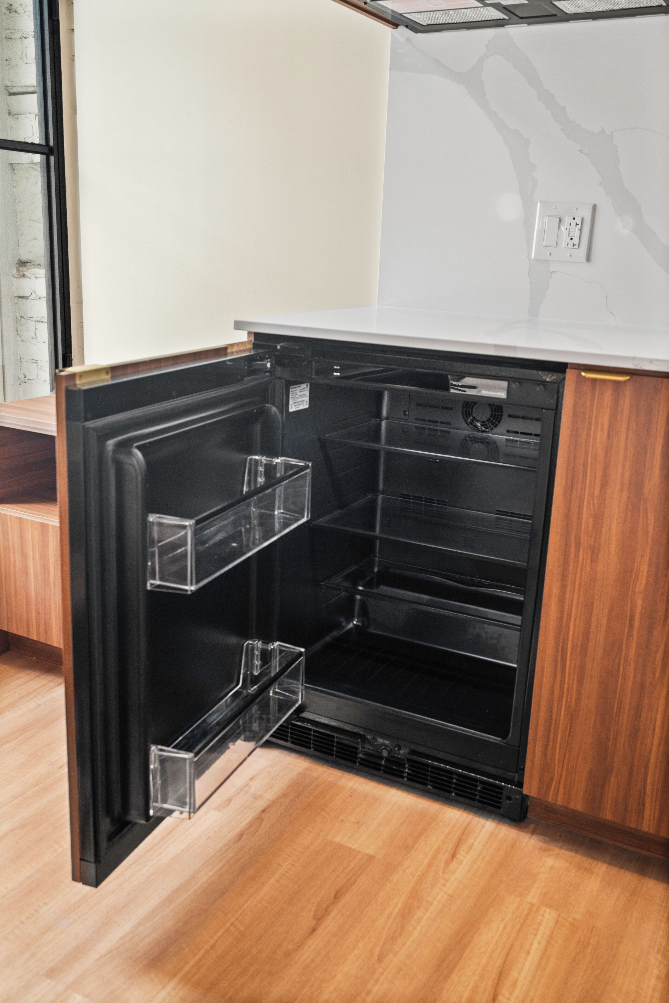 A compact refrigerator with a transparent door, built into a wooden cabinet with a white countertop above.