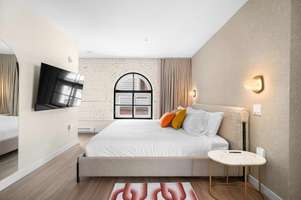 Un dormitorio moderno con una cama grande, un televisor de pantalla plana montado en la pared, una mesita de noche pequeña y una ventana con cortinas.