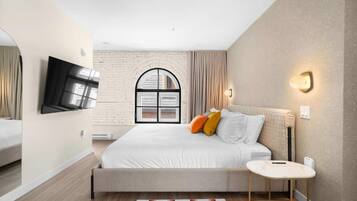 Un dormitorio moderno con una cama grande, un televisor de pantalla plana montado en la pared, una mesita de noche pequeña y una ventana con cortinas.