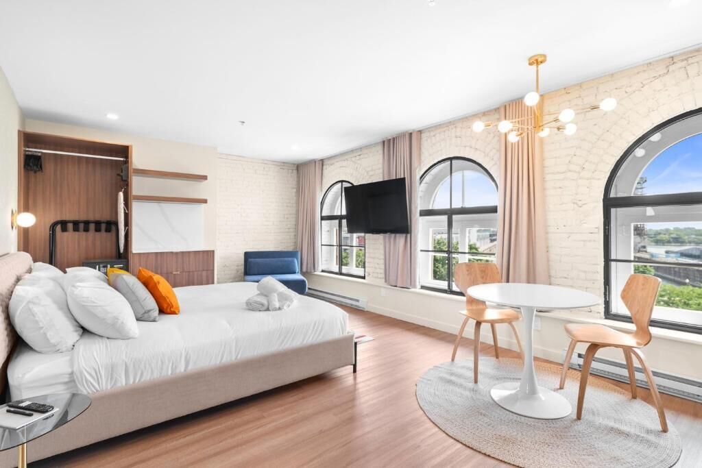 Een moderne hotelkamer met een bed, een televisie, een zithoek met stoelen en grote ramen met uitzicht op de stad.