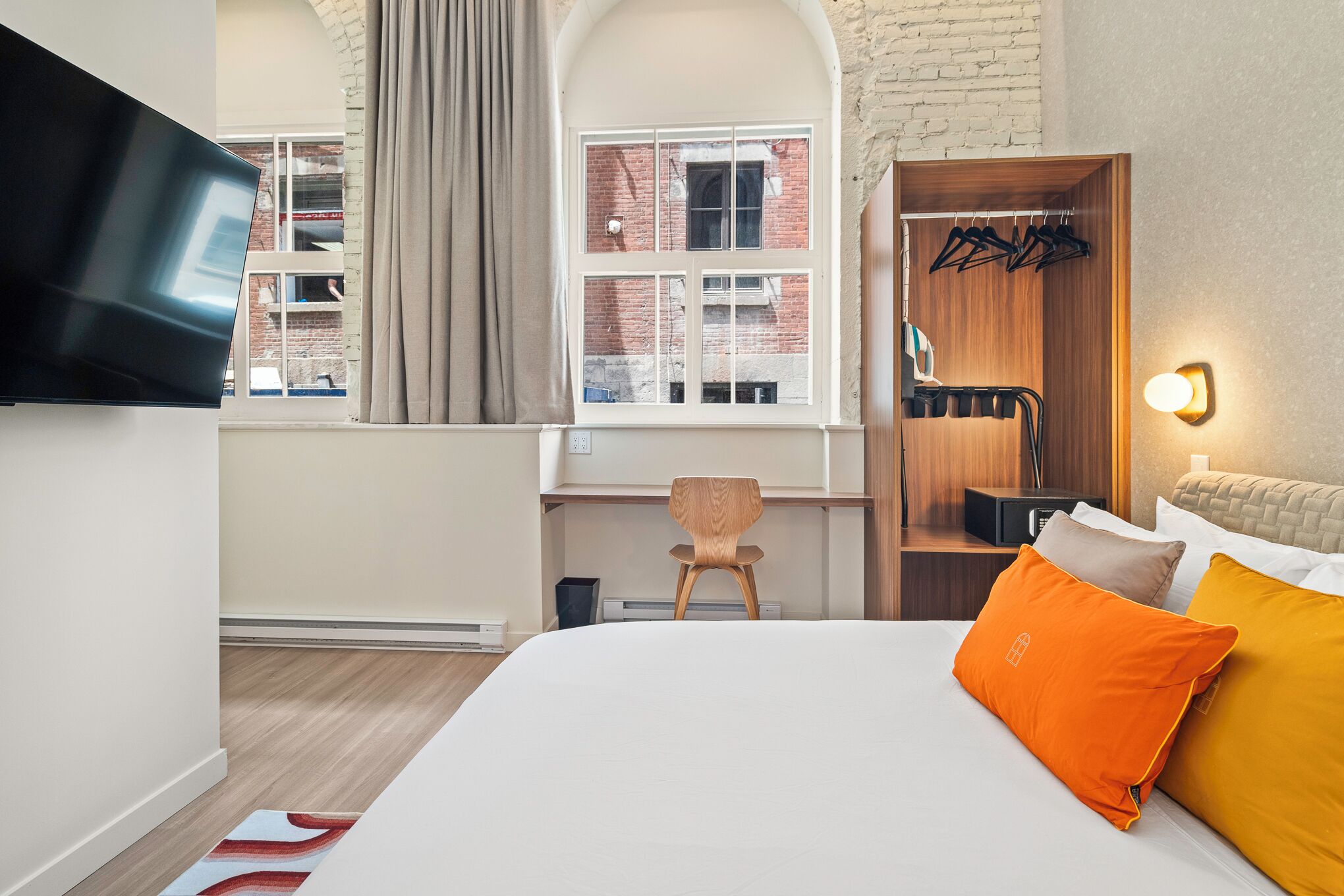 Habitación de hotel con cama, televisión montada en la pared, escritorio con silla, armario y ventana con cortinas.