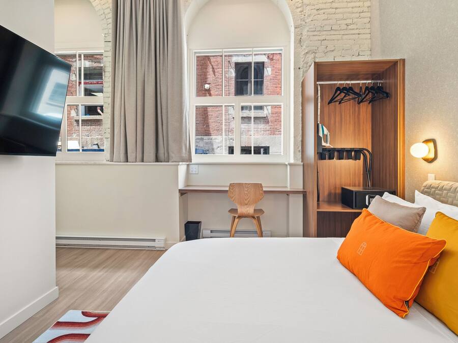 Habitación de hotel con cama, televisión montada en la pared, escritorio con silla, armario y ventana con cortinas.