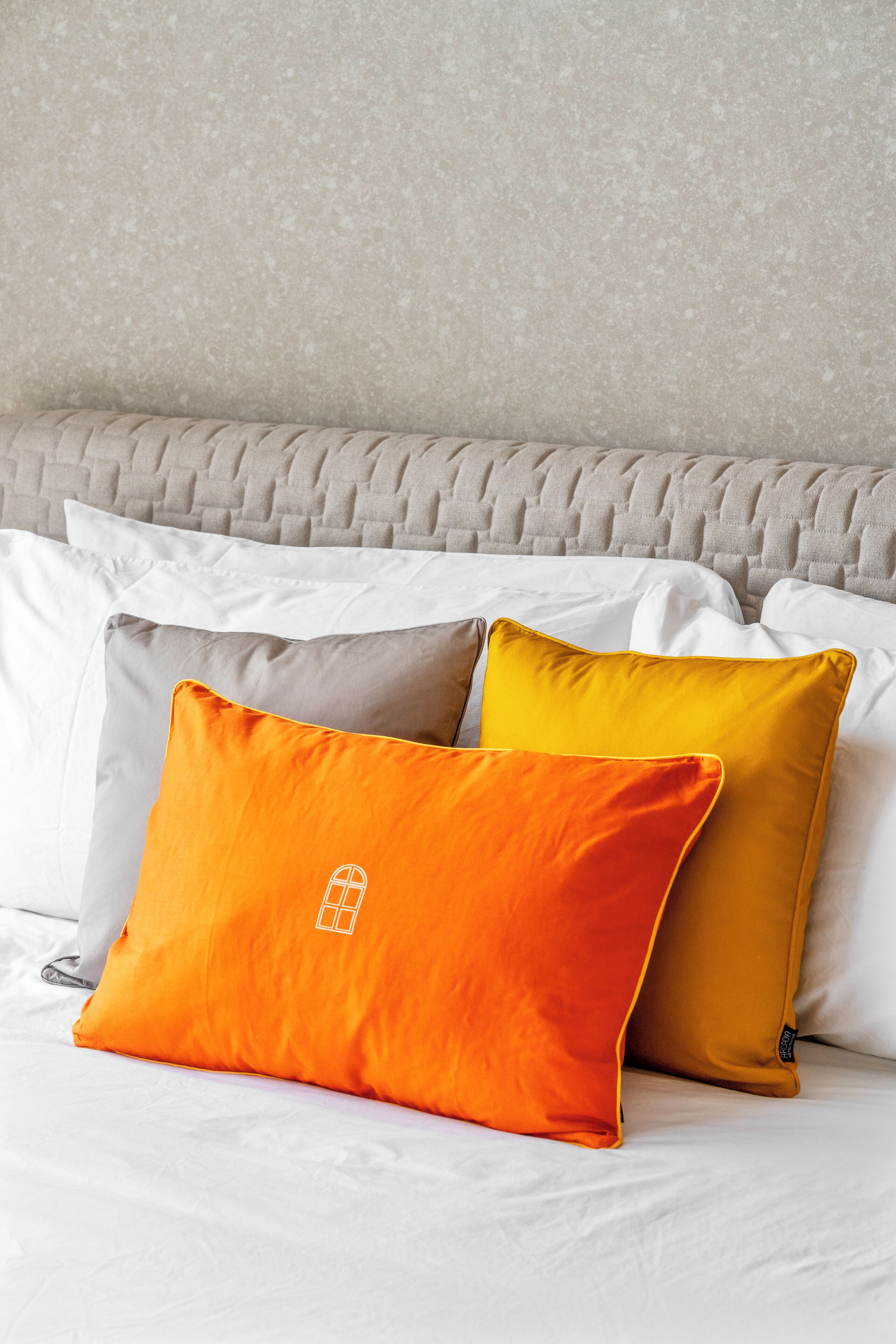 A bed with white and orange pillows, a grey headboard, and a textured wall.