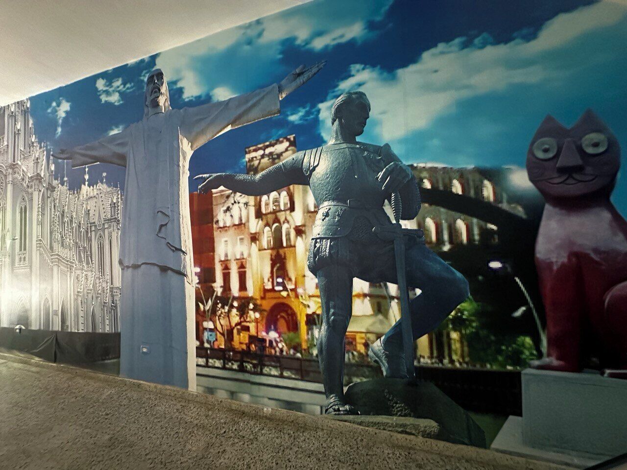 En staty av en man med utsträckta armar, en stor röd kattstaty och en väggmålning som föreställer en stadsbild.