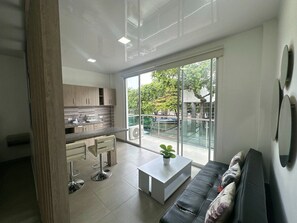A modern kitchen with a large window, a dining area with a white table and chairs, and a living room with a sofa and a plant.
