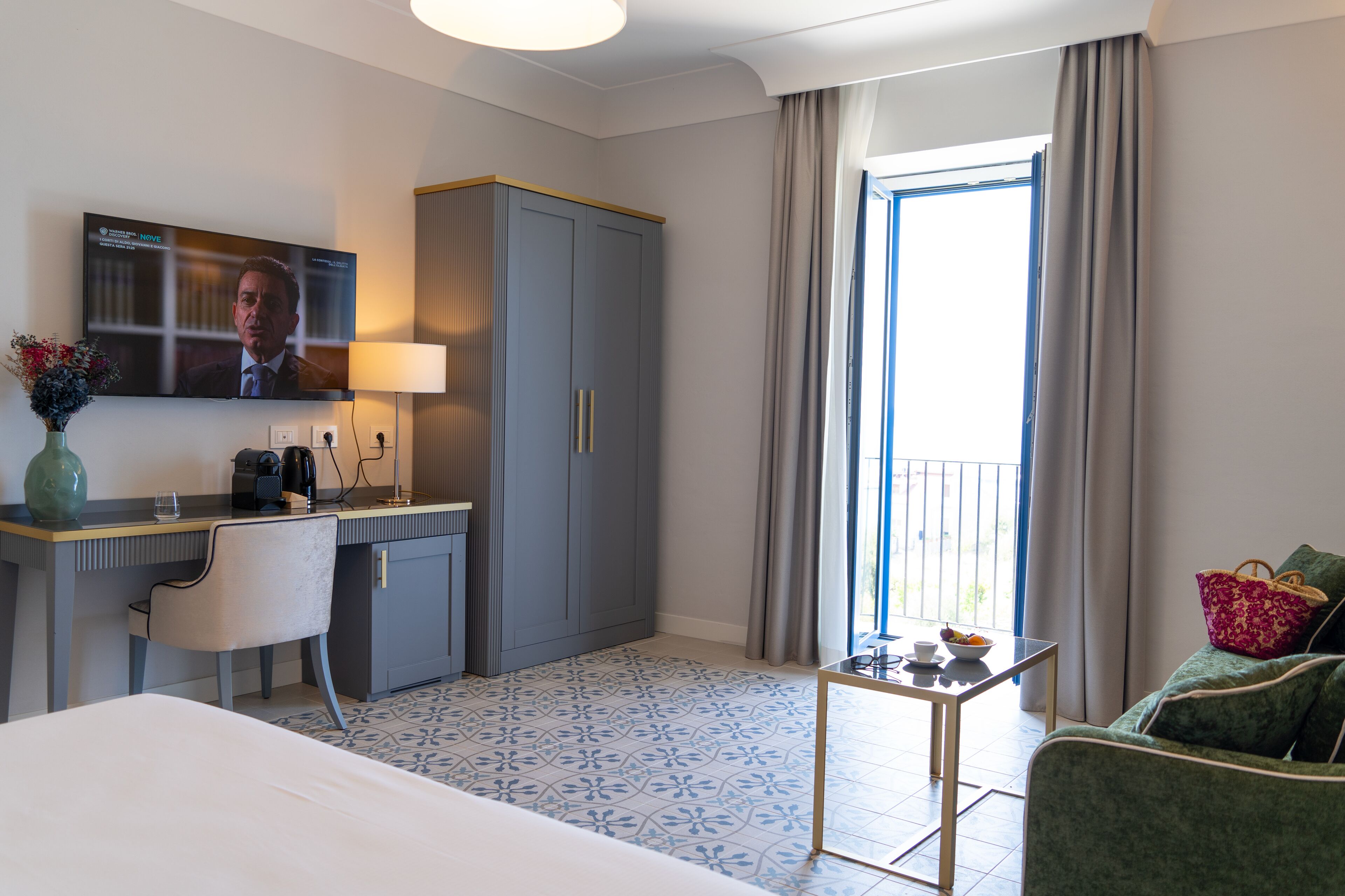 A hotel room with a bed, a desk with a chair, a television, a wardrobe, a small table with a lamp, and a view of the outdoors through a window.