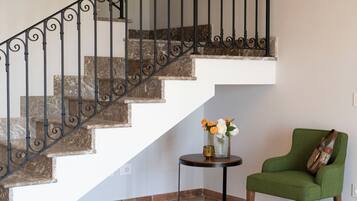 Eine Treppe mit Marmorverkleidung und schwarzem Eisengeländer, ein kleiner runder Tisch mit Blumen in einer Vase und ein mit grünem Stoff gepolsterter Sessel.