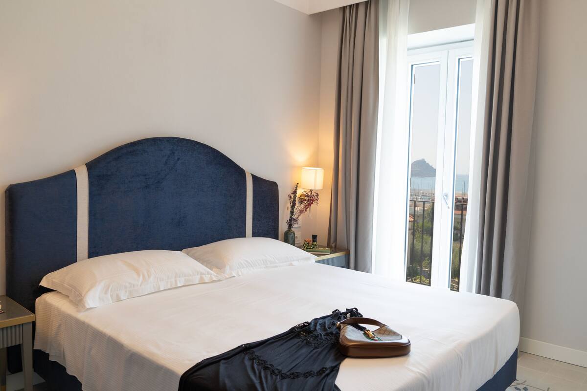 Habitación de hotel con una cama grande, una mesita de noche, una lámpara y vistas al exterior por una ventana.