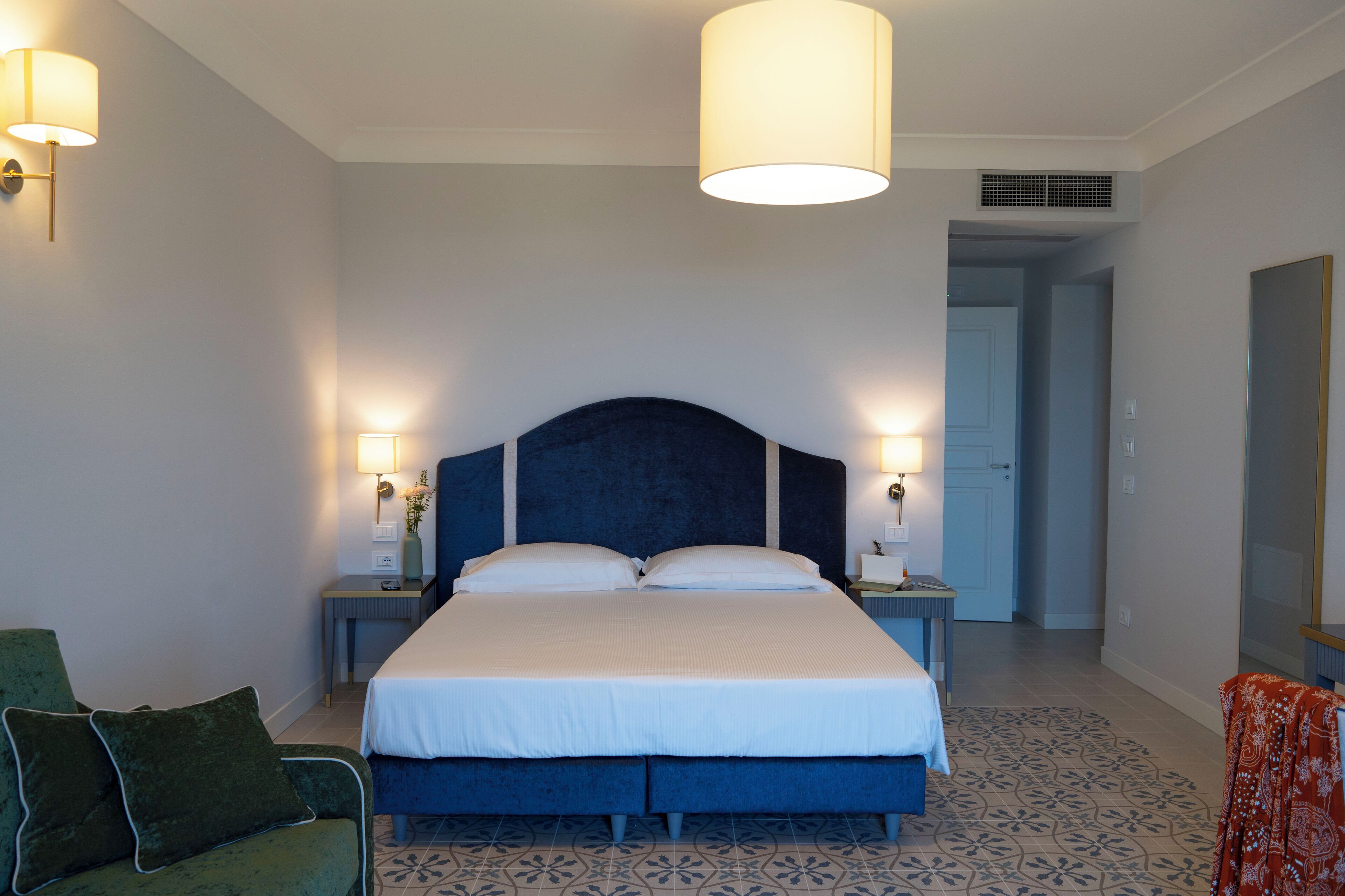 Ein Schlafzimmer mit einem großen Bett, zwei Nachttischen, einem Beistelltisch, einem grünen Sessel und einem gemusterten Boden.