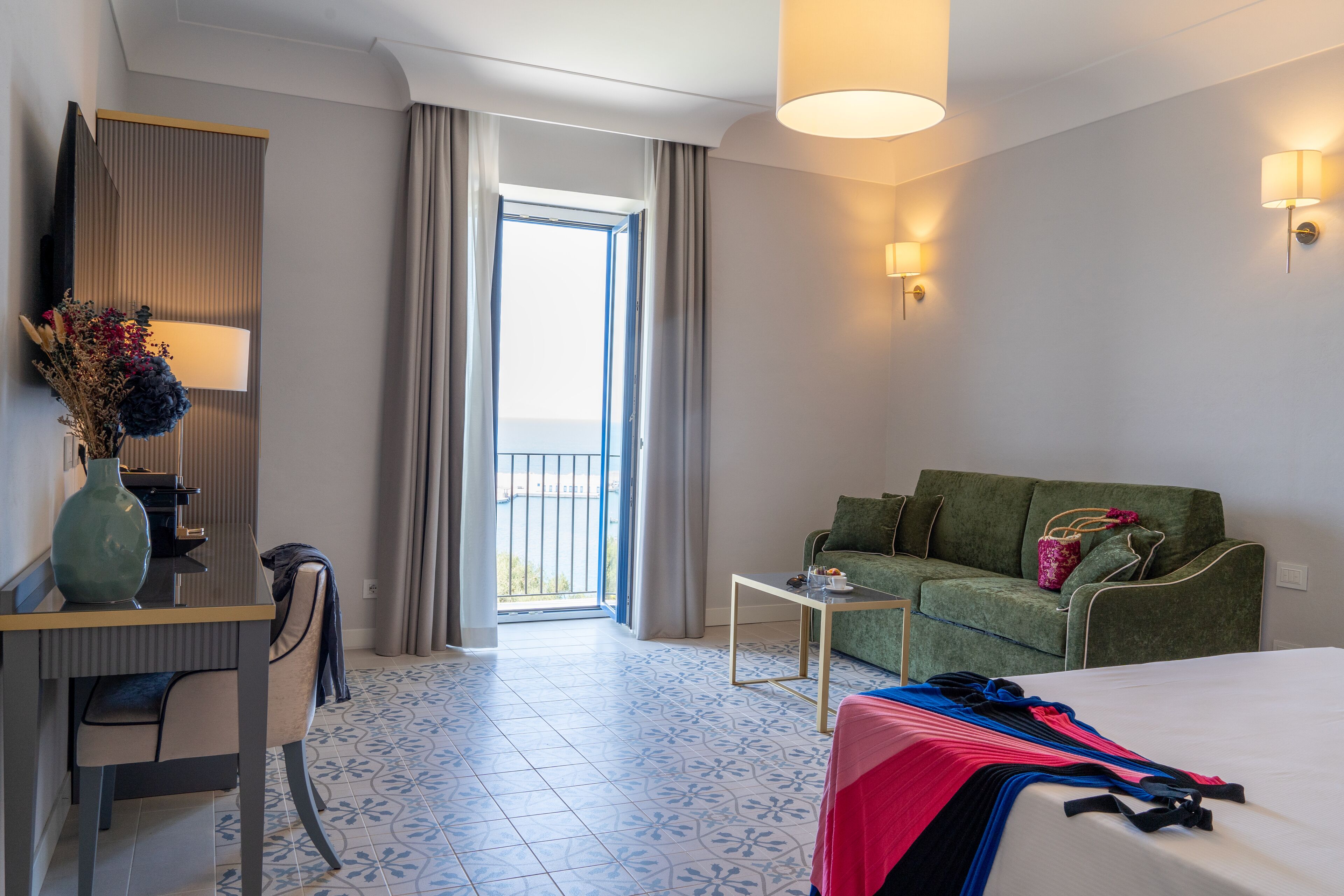 Une chambre d’hôtel avec un lit, un canapé vert, un bureau avec une lampe et une vue sur l’extérieur grâce à une grande fenêtre.