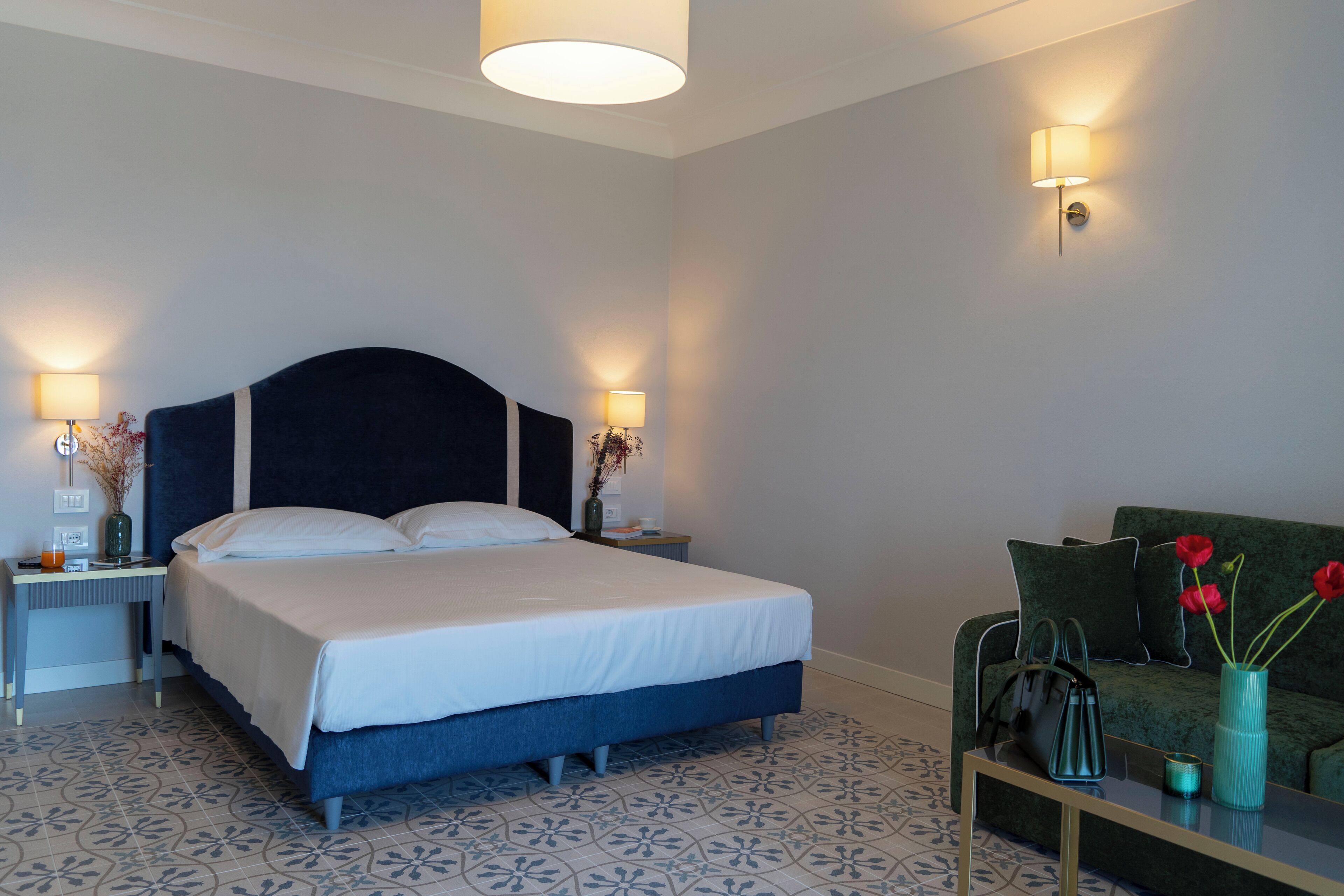 Une chambre bien rangée, avec un lit, des tables de chevet, un canapé vert et un vase rempli de fleurs.
