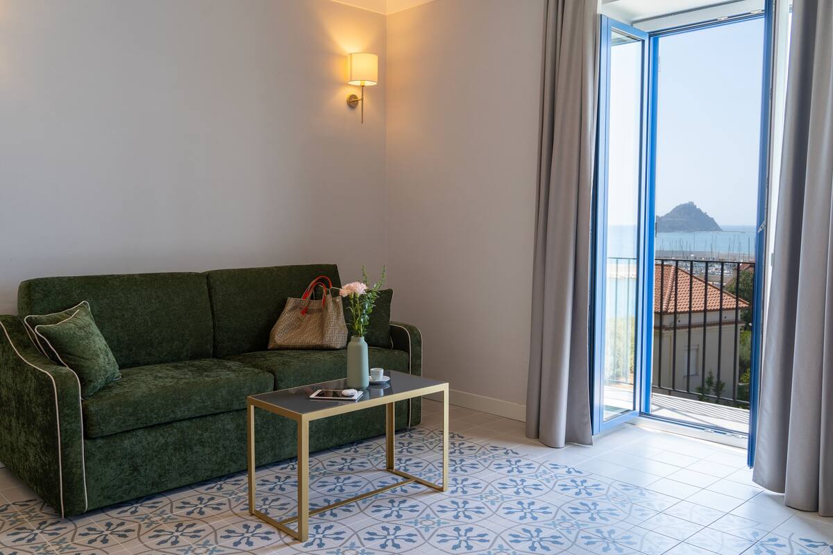 Un sofá verde, una mesita con un florero y un bolso, y vista al mar desde un balcón.