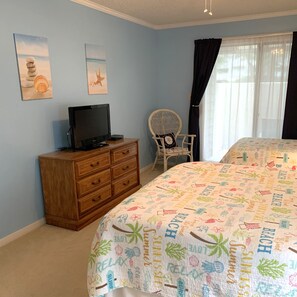 Een slaapkamer met een bed, een houten dressoir, een televisie, een rieten stoel en twee kunstwerken aan de muur.