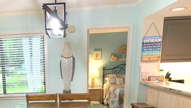 A dining area with a glass-top table and wooden chairs, a bed with a patterned bedspread, and a kitchen area with a countertop and stools. - Beach Retreat 3BR Ground Floor Condo in Kingston Plantation! (Myrtle Beach)