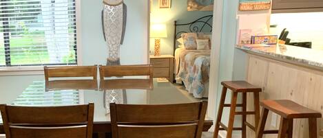 A dining area with a glass-top table and wooden chairs, a bed with a patterned bedspread, and a kitchen area with a countertop and stools.