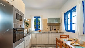 A compact kitchen with a refrigerator, oven, and sink. There is a dining table with chairs and a window with blue shutters.