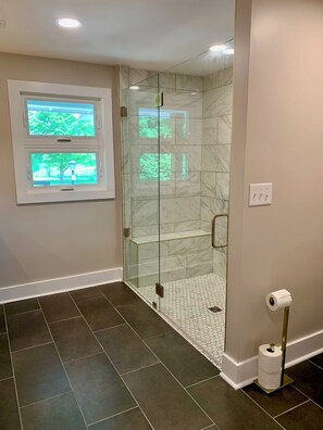 A modern bathroom with a glass-enclosed shower, dark tiled flooring, a toilet paper holder, and a window with a view of greenery.