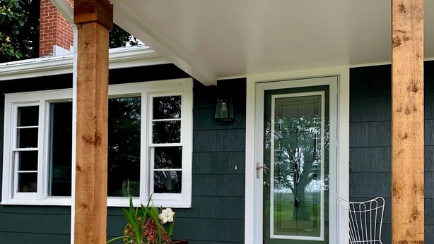 A covered porch with wooden columns, a potted plant, a chair, and a door with a glass panel.