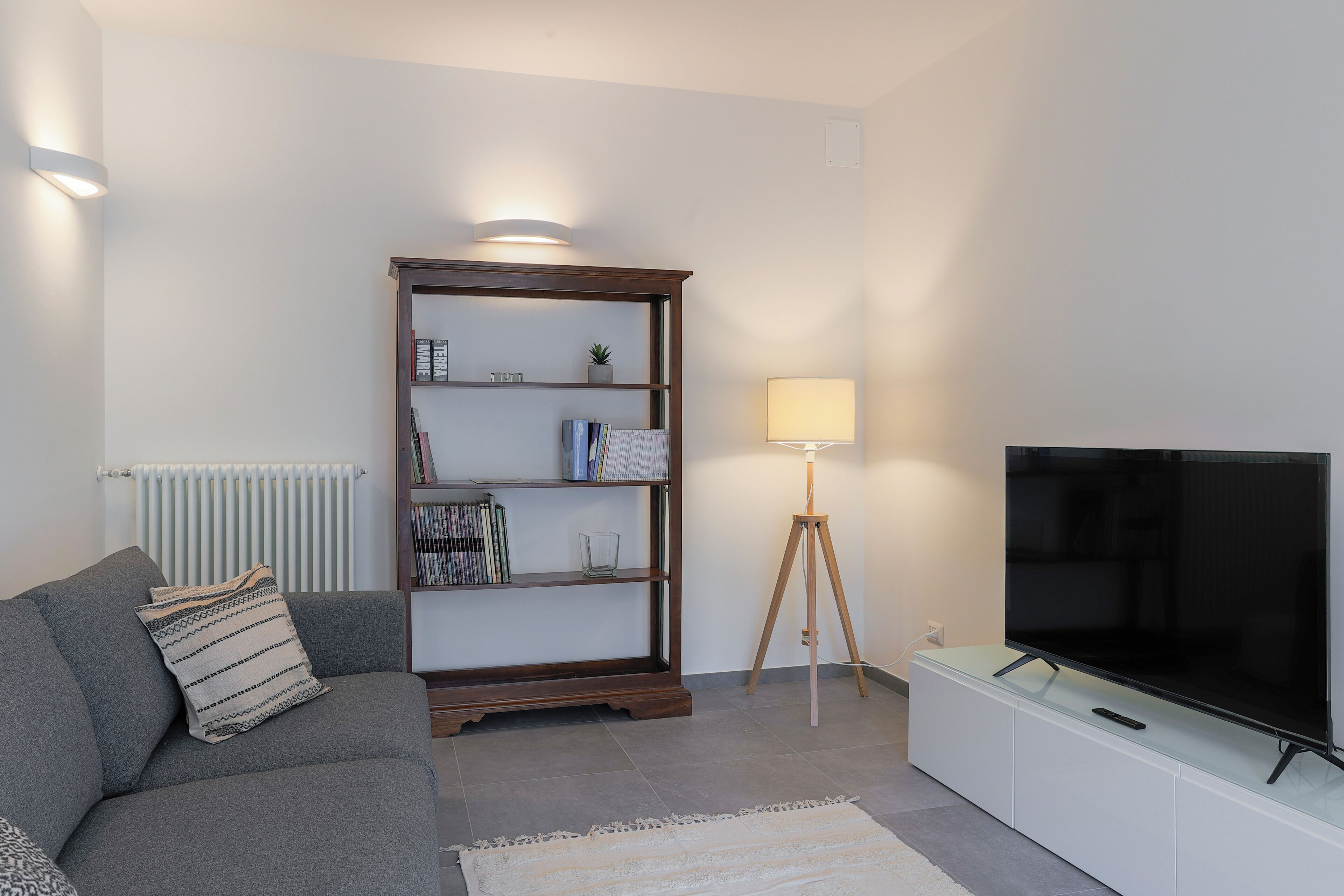 Una sala de estar moderna con un sofá gris, una estantería de madera, una lámpara de pie y un televisor de pantalla plana sobre un mueble blanco.