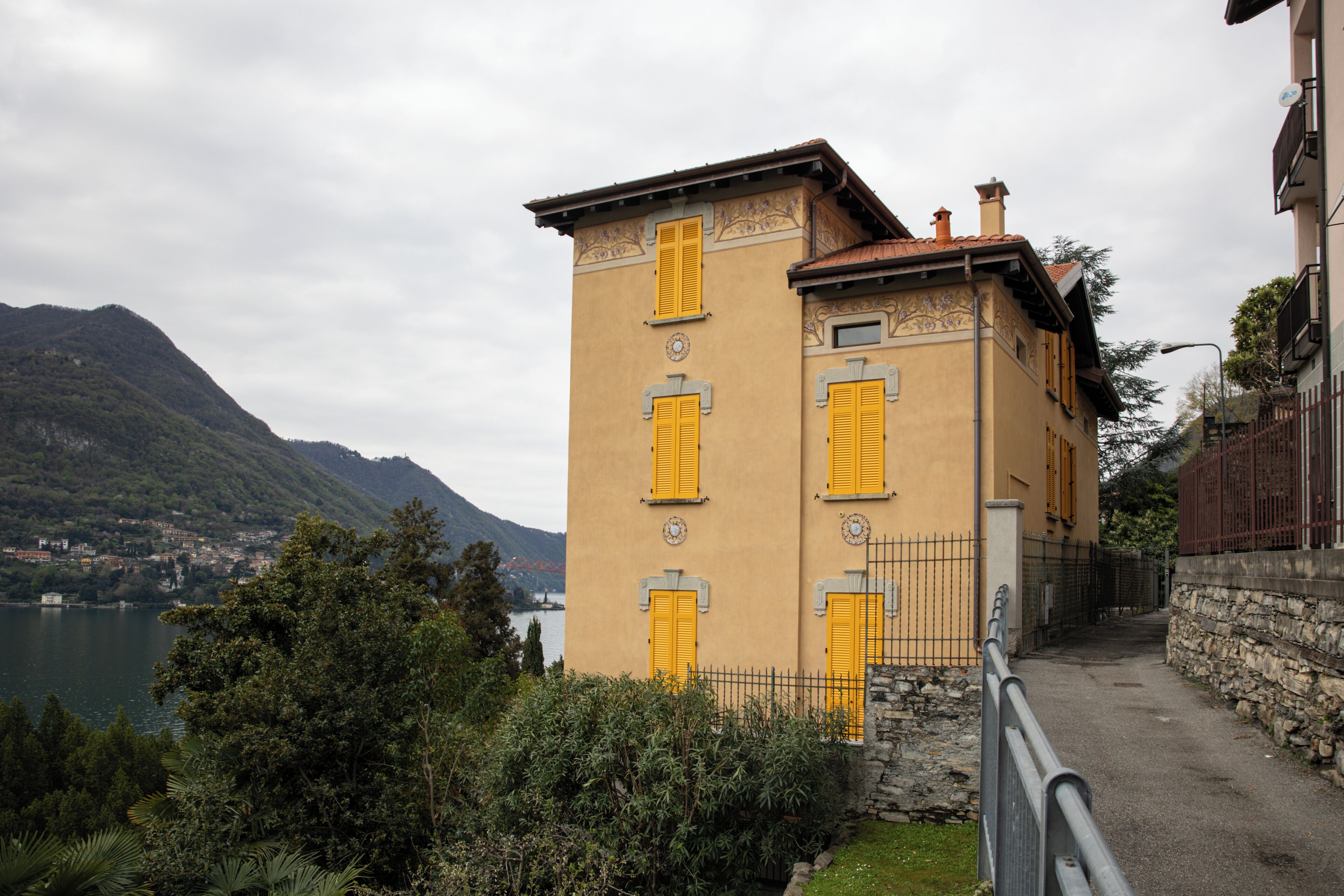 Un edificio con contraventanas amarillas, un muro de piedra y vistas a un lago y montañas.