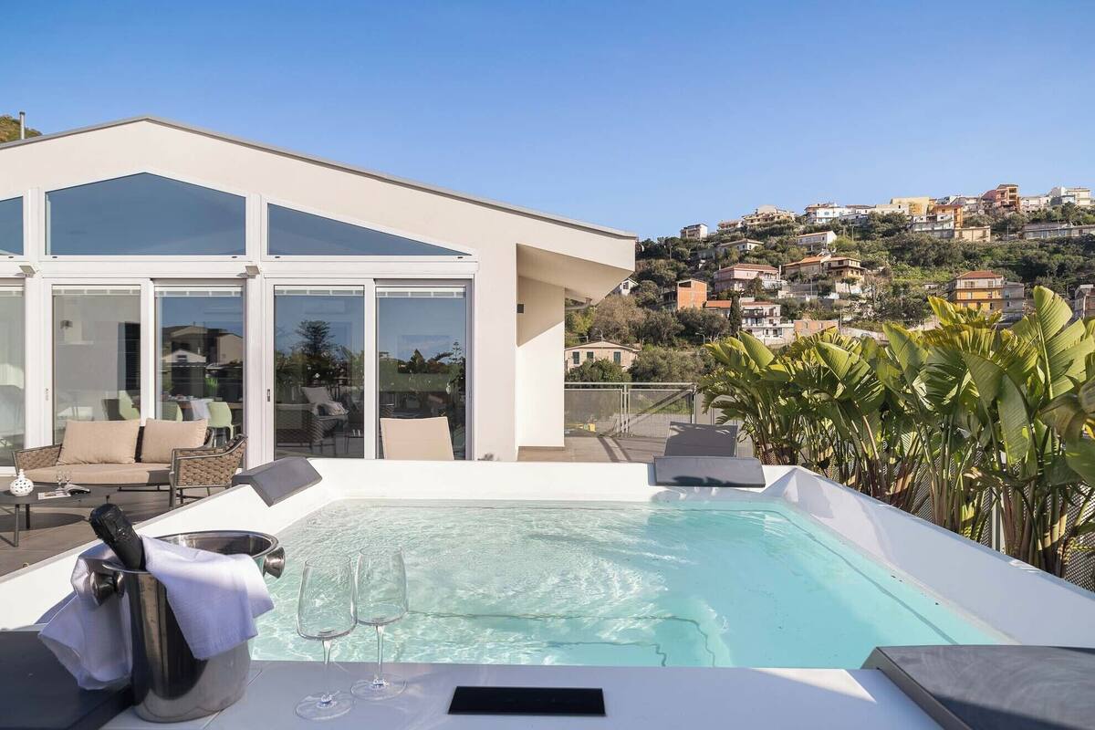 Un área de piscina en la azotea con un salón con paredes de vidrio, un jacuzzi y vistas a un pueblo en la ladera.
