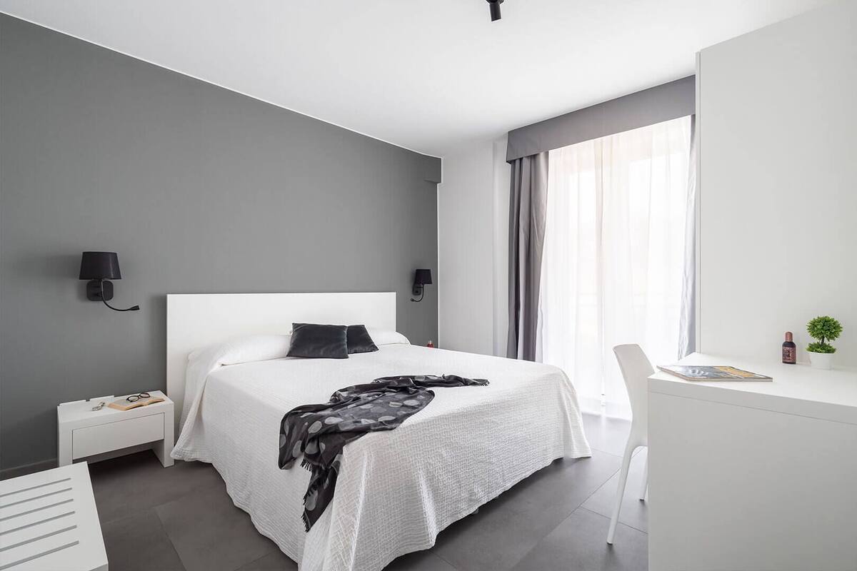 Un dormitorio moderno con una cama blanca, paredes grises y un amplio ventanal con cortinas.