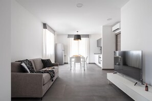Un salon moderne avec un canapé gris, une table à manger avec des chaises, un coin cuisine et un téléviseur à écran plat fixé sur un meuble blanc.