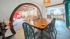 A spacious living area with a wooden dining table, teal chairs, and a large arched doorway leading to a kitchen. - The Old Barn Near St Ives - Rustic Charm & Space (St Ives)