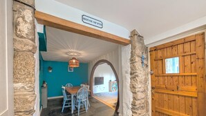 A dining area with a wooden table and blue chairs, a stone pillar, and a wooden door with a sign above it. - The Old Barn Near St Ives - Rustic Charm & Space (St Ives)