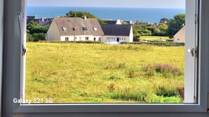 Uitzicht door een raam op een huis met een grasveld, een wit gebouw en de zee op de achtergrond.