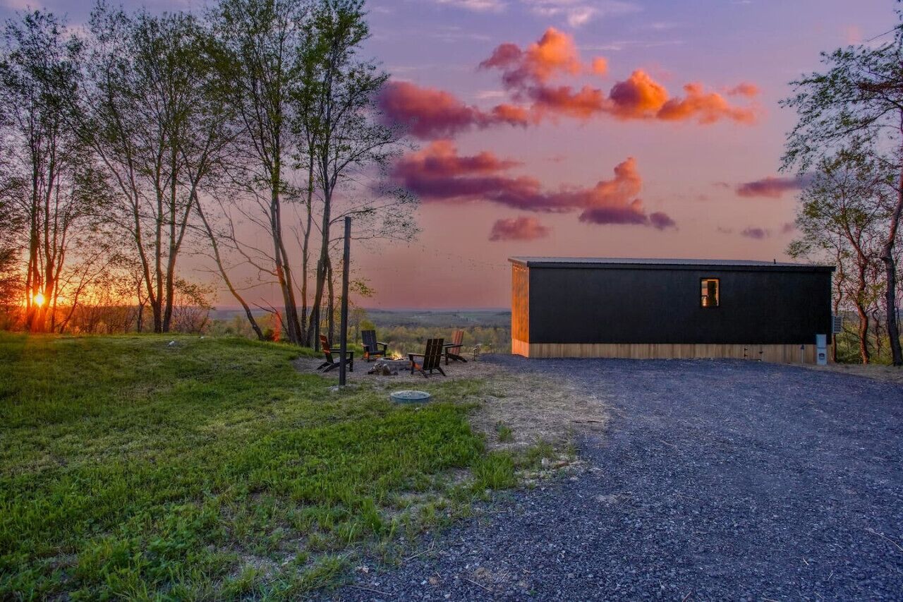 Een rustieke hut met een grindpad, omringd door bomen en uitzicht op een zonsondergang.