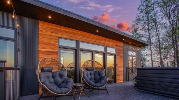 A modern building with large windows, a wooden deck, and two wicker chairs. - Skyline Serenity | Hot Tub, Panoramic Views, King Bed in PA Wilds (Brookville)