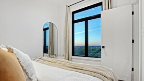 A bedroom with a large window, a bed with white bedding, a mirror, and a door. - Skyline Serenity | Hot Tub, Panoramic Views, King Bed in PA Wilds (Brookville)