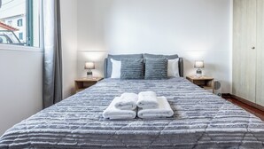 A neatly made bed with folded towels, two bedside lamps, and a view of the outside through a window. - Cozy Seaside Cottage | Ocean Views & Near Beach (Caniço)