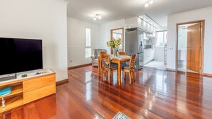A spacious living area with a wooden floor, a dining table with chairs, a flat-screen TV on a wooden stand, and a kitchen in the background. - Cozy Seaside Cottage | Ocean Views & Near Beach (Caniço)