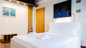 Une chambre d’hôtel avec un grand lit, du linge de lit blanc, une porte en bois, un téléviseur fixé au mur et des œuvres d’art accrochées aux murs.