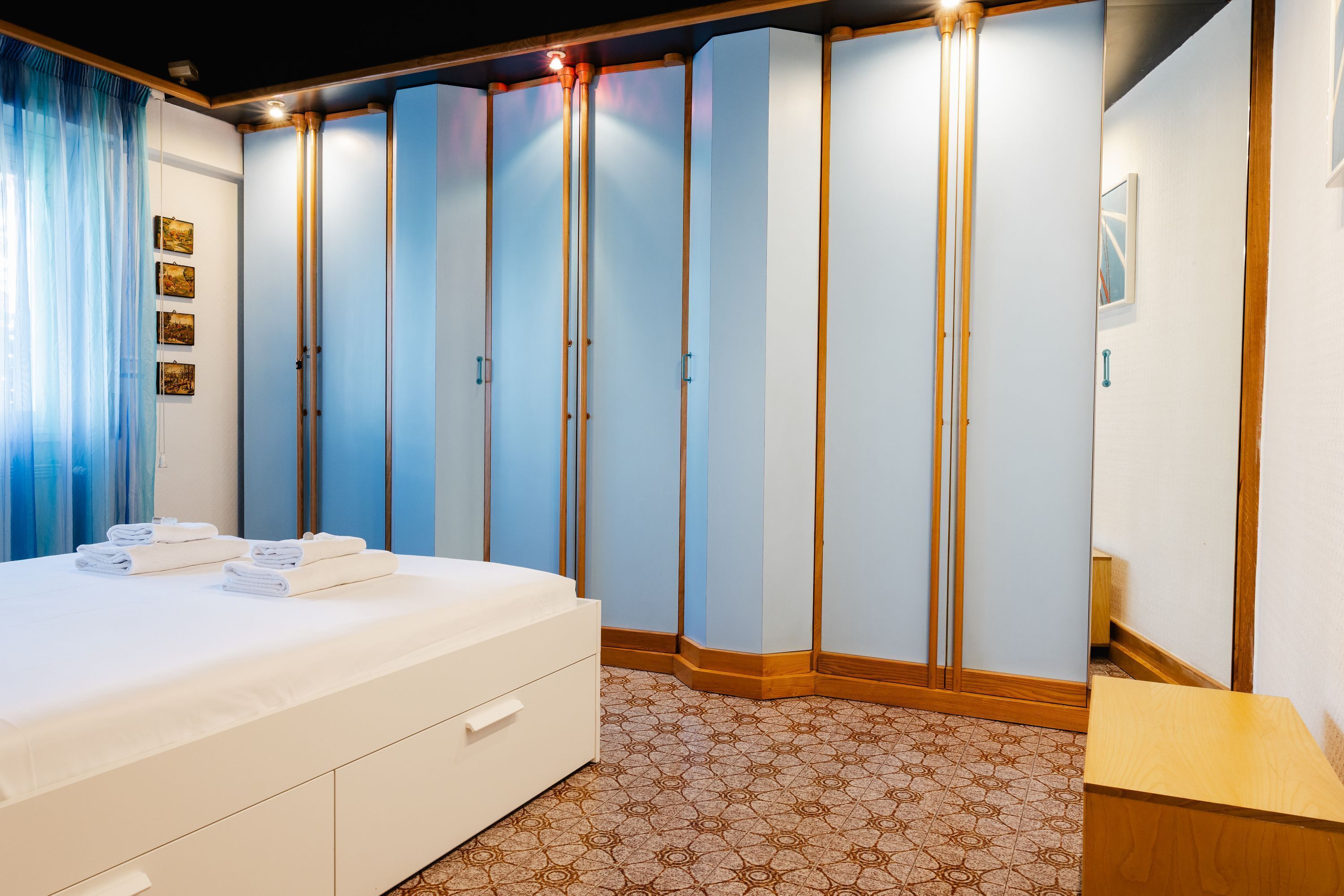 A hotel room with a bed, a wooden nightstand, a wardrobe, and a patterned floor.