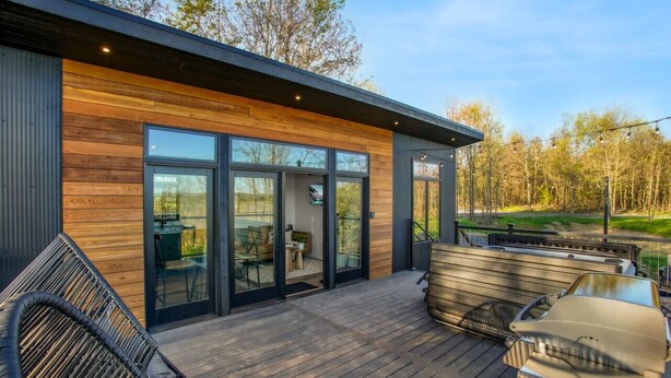Une maison moderne avec une façade en bois, de grandes portes vitrées, une terrasse avec des meubles d’extérieur et un barbecue.