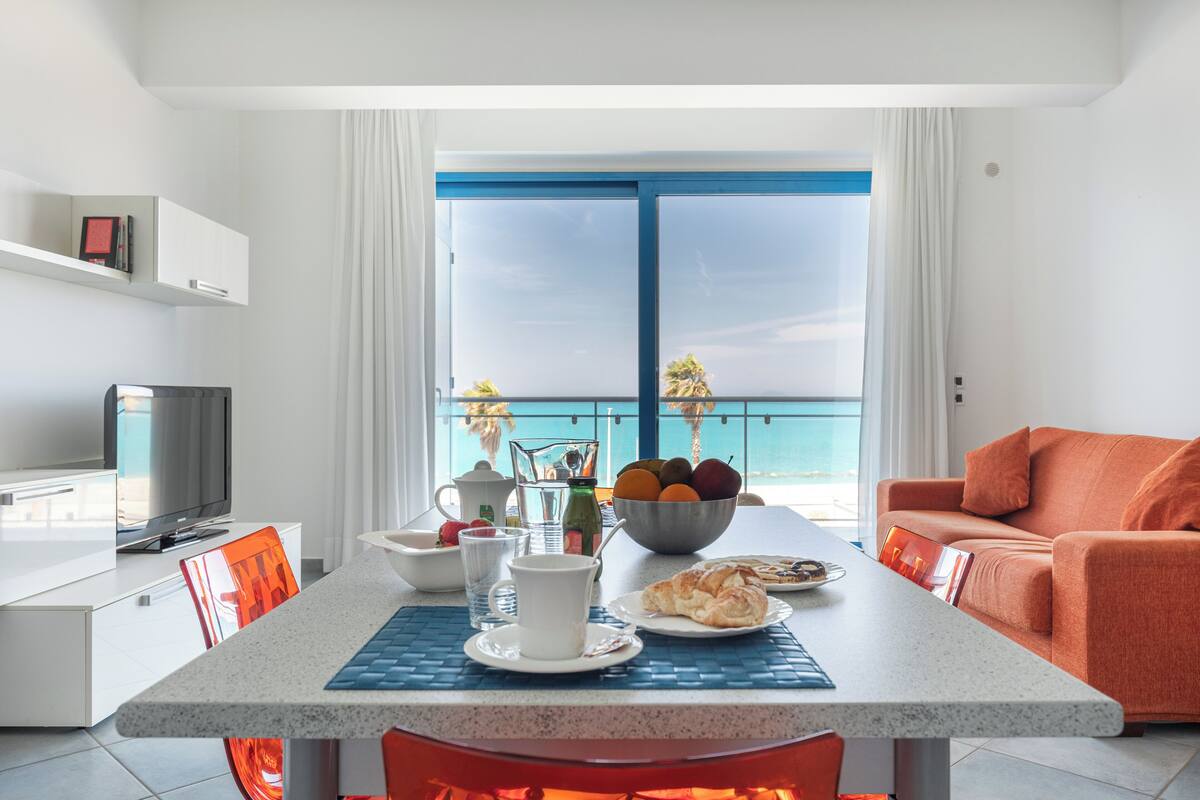Une chambre d’hôtel moderne dotée d’un coin repas, d’une télévision et d’un balcon offrant une vue sur la mer.