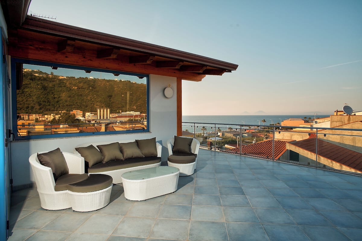 Une terrasse sur le toit, avec un ensemble de meubles de jardin en osier blanc, une table basse en verre et une vue sur la mer et les collines lointaines.