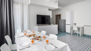 Un comedor moderno con una mesa blanca dispuesta para el desayuno, una cocina al fondo y un amplio ventanal con cortinas.